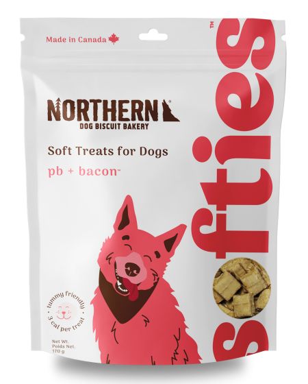 A bag of Northern Dog Biscuit Bakery soft treats labeled "pb + bacon" and "Made in Canada." The package features a pink, cartoon-style dog with a smile, and includes an image of the treats.