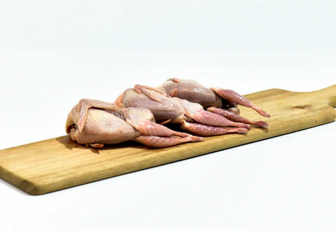 Four raw, whole quails are arranged on a wooden cutting board against a white background.