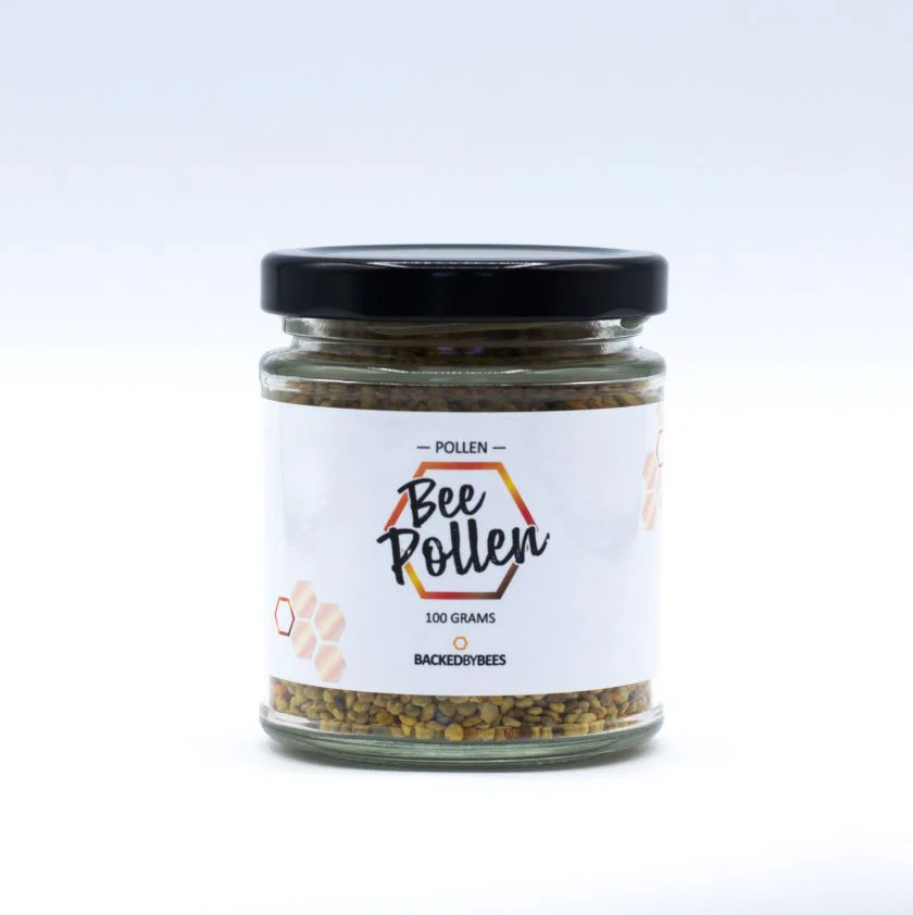 A glass jar with a black lid containing bee pollen granules. The white label reads "Bee Pollen" with a honeycomb design and states "100 grams" and "BACKEDBYBEES." The jar is set against a plain white background.