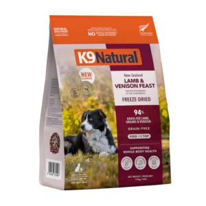 A bag of K9 Natural freeze-dried dog food, Lamb & Venison Feast flavor, featuring a photo of a black and white dog lying on grass. The packaging highlights grain-free ingredients and whole-body health support.