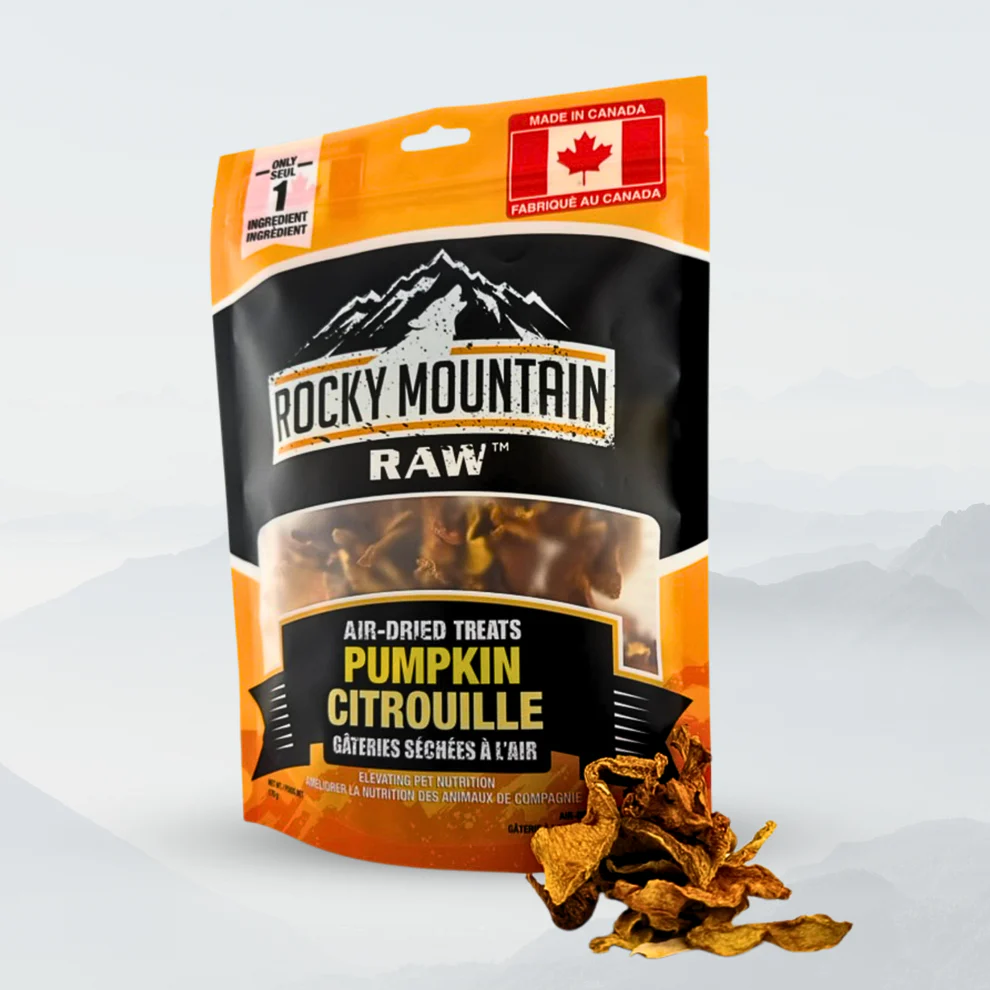 A bag of Rocky Mountain Raw air-dried pumpkin treats stands upright, displaying orange, black, and white packaging with a mountain logo and Canadian flag. Dried pumpkin pieces are scattered in front of the bag.