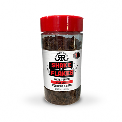 A clear plastic container with a red lid labeled "Shake A Flakes Meal Topper Beef Liver for Dogs & Cats" by Raised Right, filled with dark brown flakes.