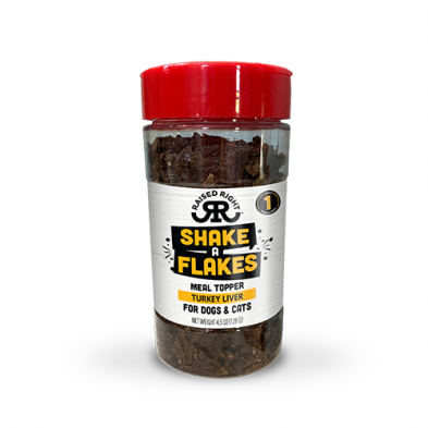 A clear plastic jar with a red lid containing "Shake A Flakes" turkey liver meal topper for dogs and cats by Raised Right. The label is white with black and yellow text and a small turkey icon.