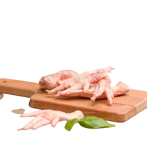 A wooden cutting board with several raw chicken feet stacked on it. In front of the board, there are a few more chicken feet and a sprig of fresh green leaves. The background is plain and out of focus.