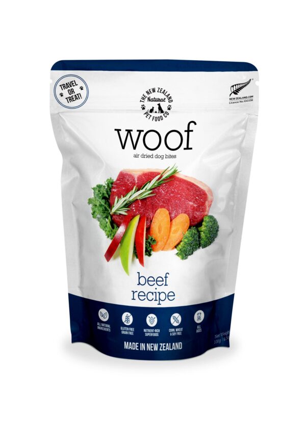 A blue and white package of Woof Air Dried Beef dog Bites 100 g showcases a photo of raw beef slices, green herbs, carrots, apples, and broccoli. The packaging emphasizes its New Zealand origin and features icons highlighting its unique qualities.