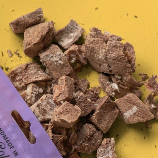 Chunks of Wild Vibes Kangaroo & Bone Broth Treat 58 g for dogs and cats are scattered on a yellow surface beside a partially visible purple package in the lower left corner.