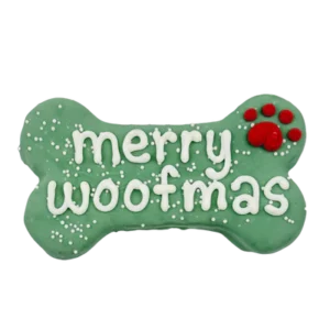 A green bone-shaped dog treat with white icing reading "merry woofmas" and a red paw print in the upper right corner, decorated with small white sprinkles.