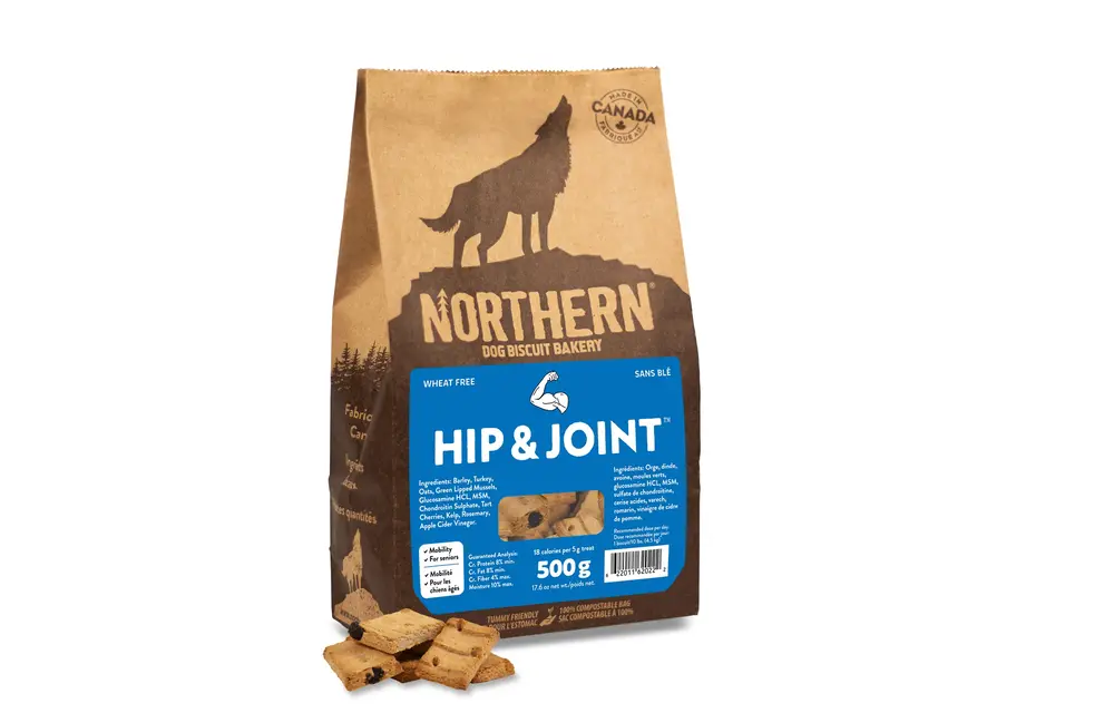 A brown paper bag of Northern Dog Biscuit Bakery Hip & Joint dog treats, featuring a wolf silhouette and a blue label. Some square treats are scattered in front of the bag. The bag states "Wheat Free" and "Made in Canada.