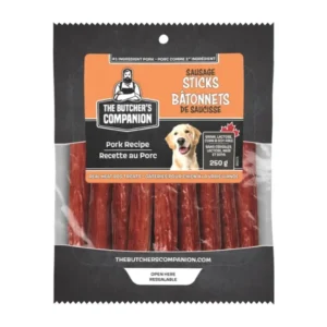 Packaging for The Butcher's Companion Sausage Sticks, featuring a cartoon butcher logo. Label highlights pork recipe with real meat dog treats, weighing 250g. A happy dog is pictured on the package. Text in English and French.