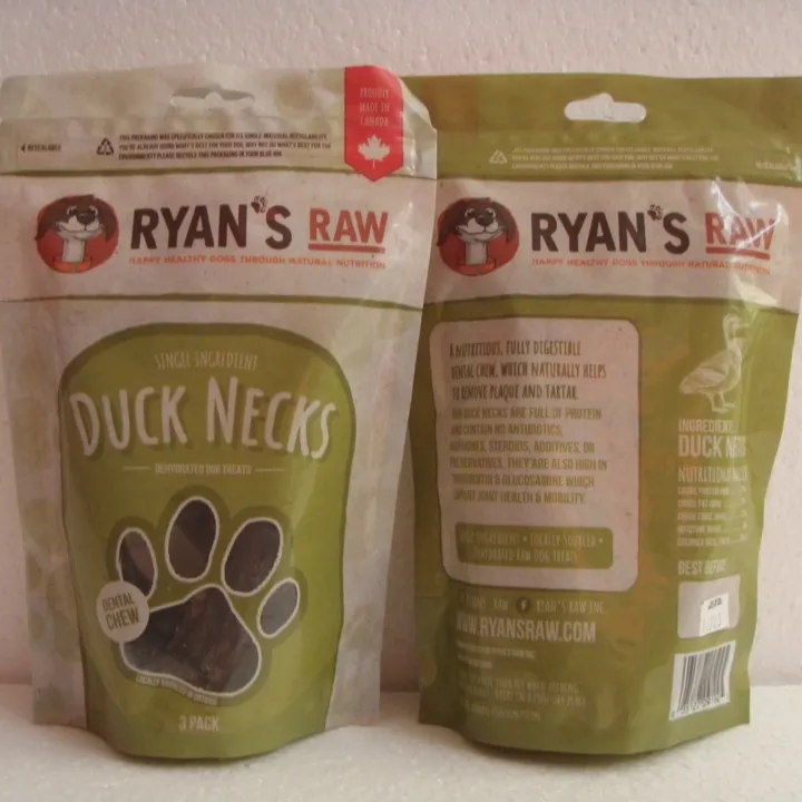 Two green and beige pouches of Ryan's Raw Duck Necks for dogs are shown. One pouch displays the front label; the other shows the back with product details and branding. Both feature a paw print and cartoon dog logo.